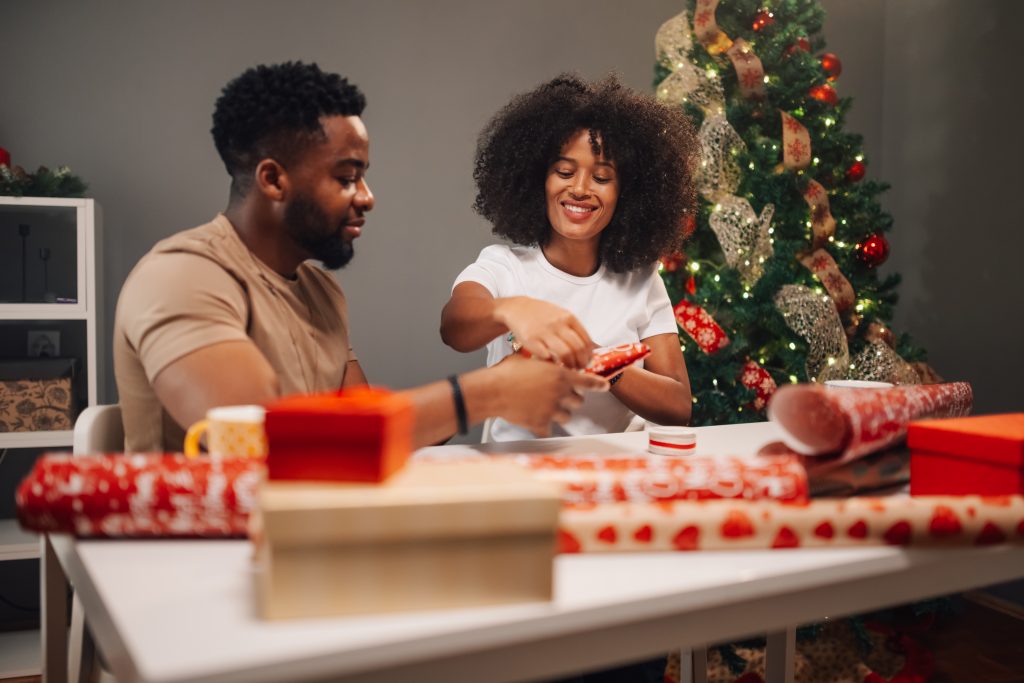Diverse couple is filled with joy, wrapping Christmas gifts in red paper, surrounded by a festively decorated tree. The image captures a cozy and cheerful atmosphere.